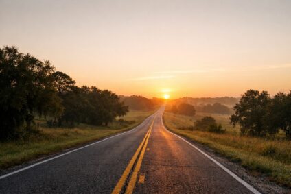 A gently rising two-lane road at sunrise, narrowing toward the horizon in soft golden light.